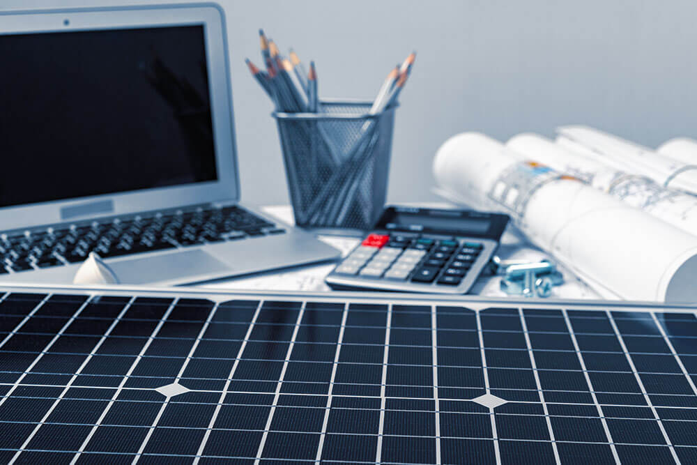 Solarzelle auf einem Schreibtisch neben Laptop, Zeichnungen, Stiften und Taschenrechner.